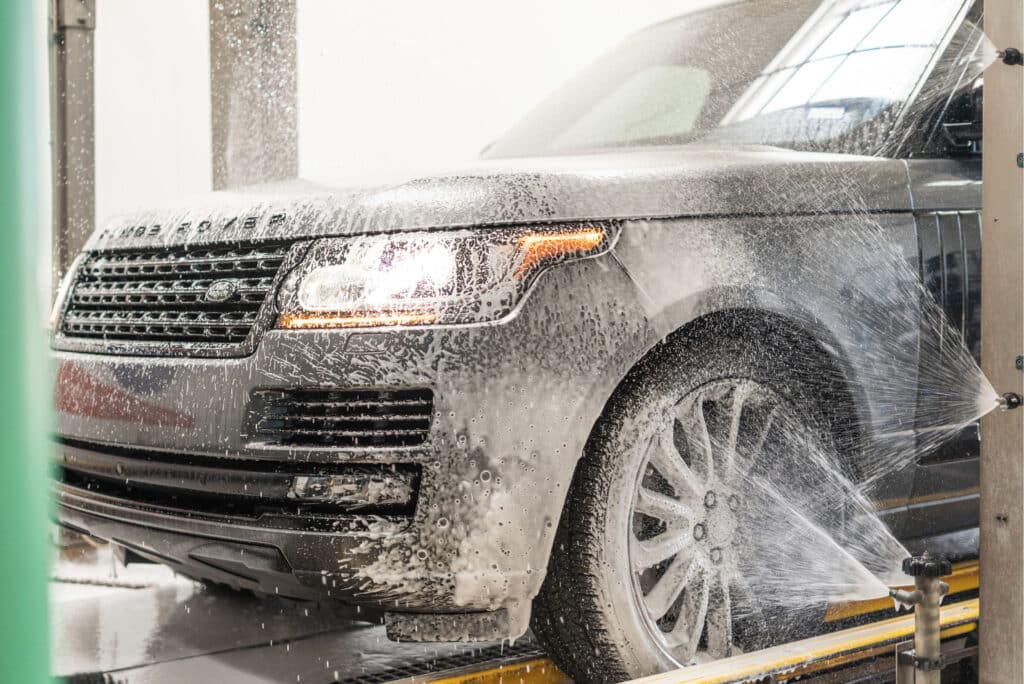 A car being thoroughly washed at a car wash, with water spraying and soap suds covering its surface.