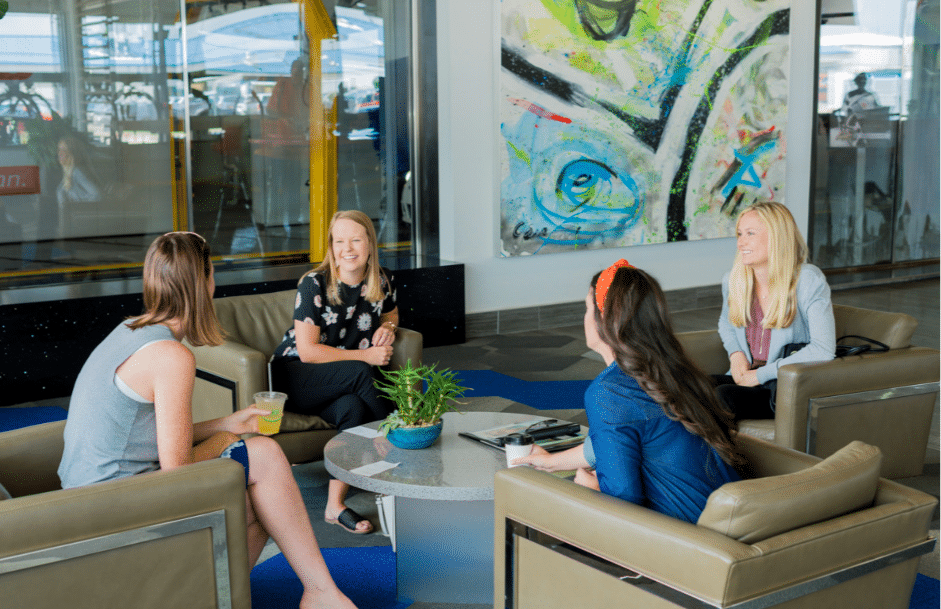 Four women seated in chairs, engaged in conversation within a modern office lobby setting.