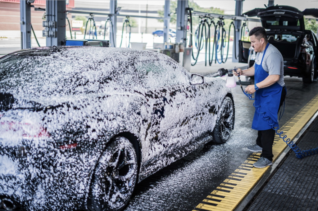 A man diligently washes a car at a car wash, surrounded by soap suds and water spray.