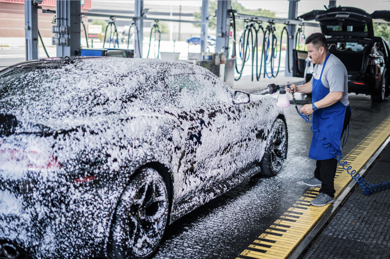 A man diligently washes a car at a car wash, surrounded by soap suds and water spray.