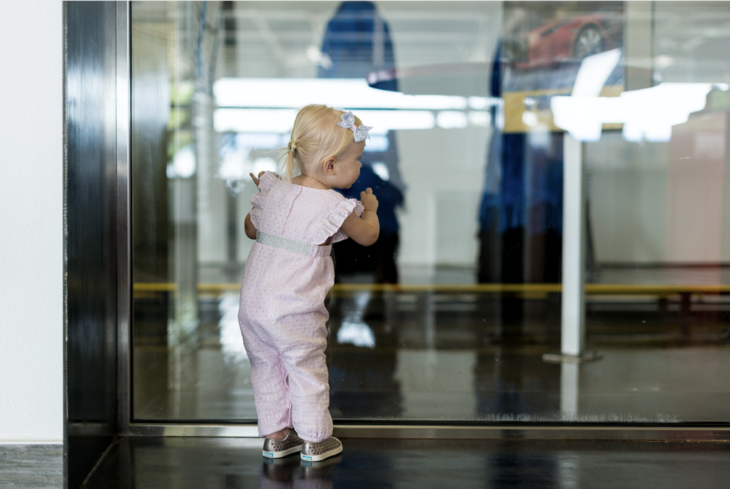 A young girl gazes curiously at a car displayed inside a modern glass building.