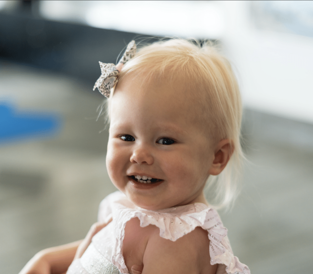 A joyful baby girl smiles brightly while seated comfortably on a chair.