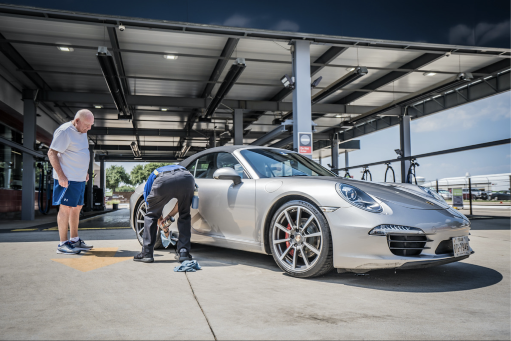Two men meticulously wash a silver Porsche car, ensuring its shine and cleanliness under bright sunlight.