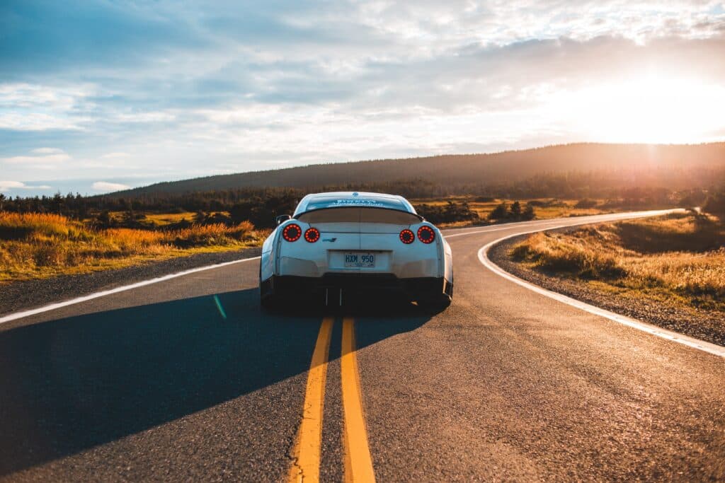 A white car travels along a road, illuminated by the warm hues of a sunset in the background.