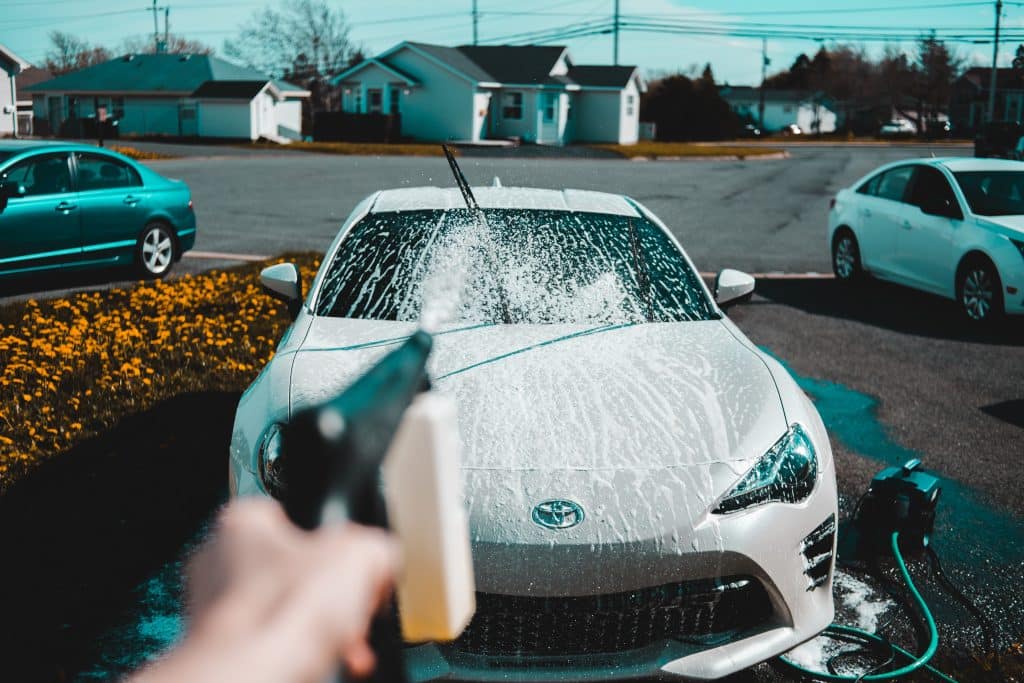 A person using a spray gun to wash a car, water spraying and soap suds visible on the vehicle's surface.