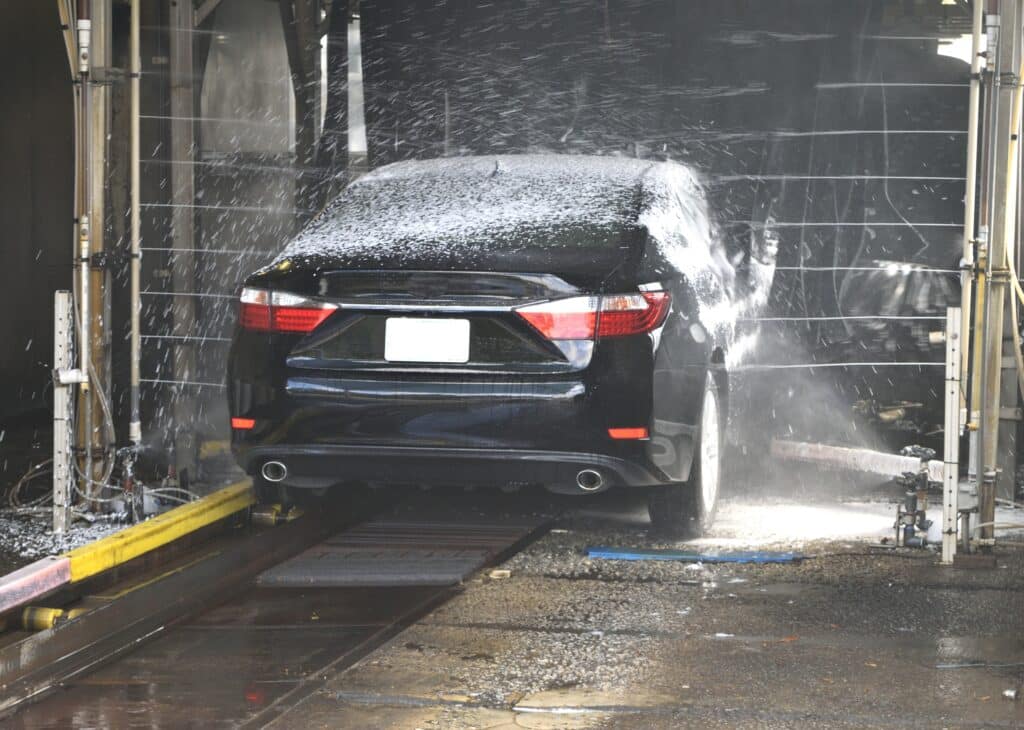 A car being thoroughly washed at a car wash, with water spraying and soap suds covering its surface.