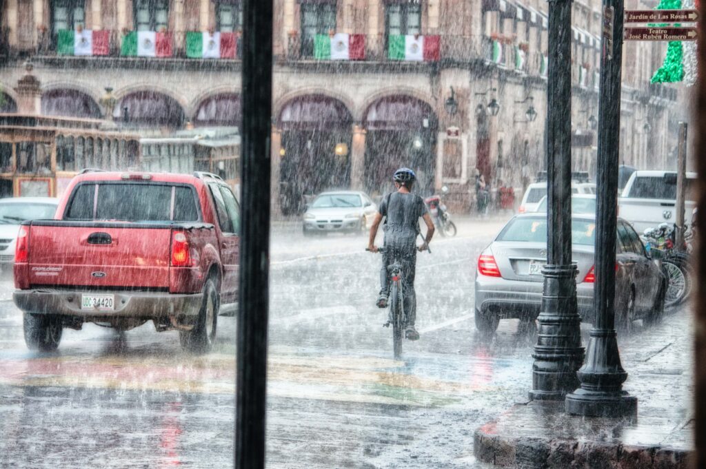 A man cycles through a rainy city street, water droplets splashing around him as he navigates the wet pavement.