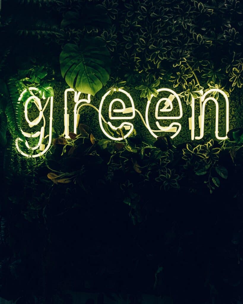 Green neon sign illuminated among lush plants, creating a vibrant and inviting atmosphere in a natural setting.