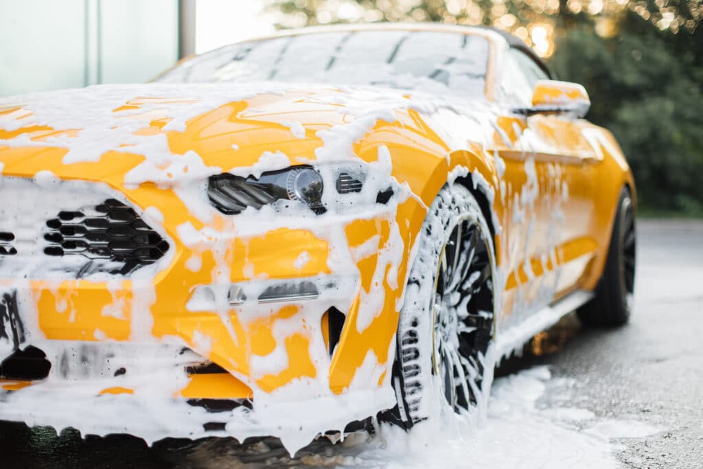 A yellow Mustang car is being washed, covered in foam, showcasing its sleek design and vibrant color.