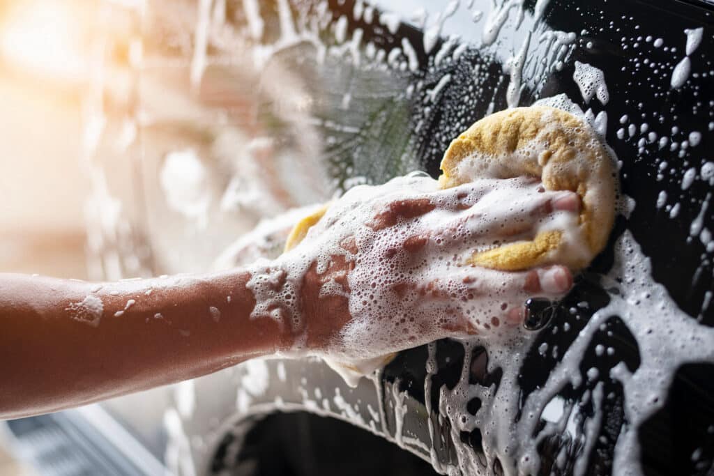 A person scrubbing a car with a sponge, ensuring it is clean and shiny under the sunlight.
