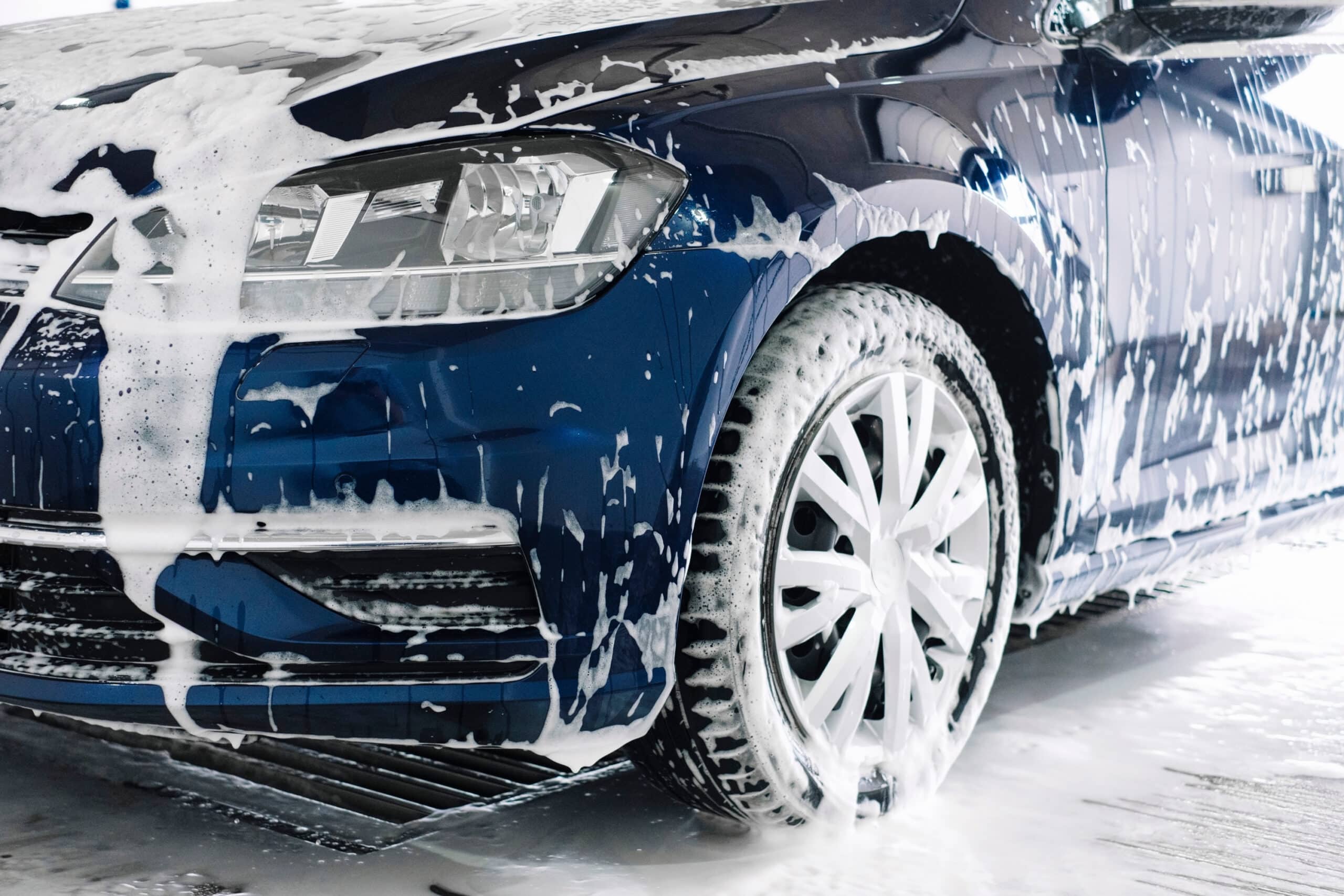 A blue car is being thoroughly washed at a car wash, with water and soap covering its surface.
